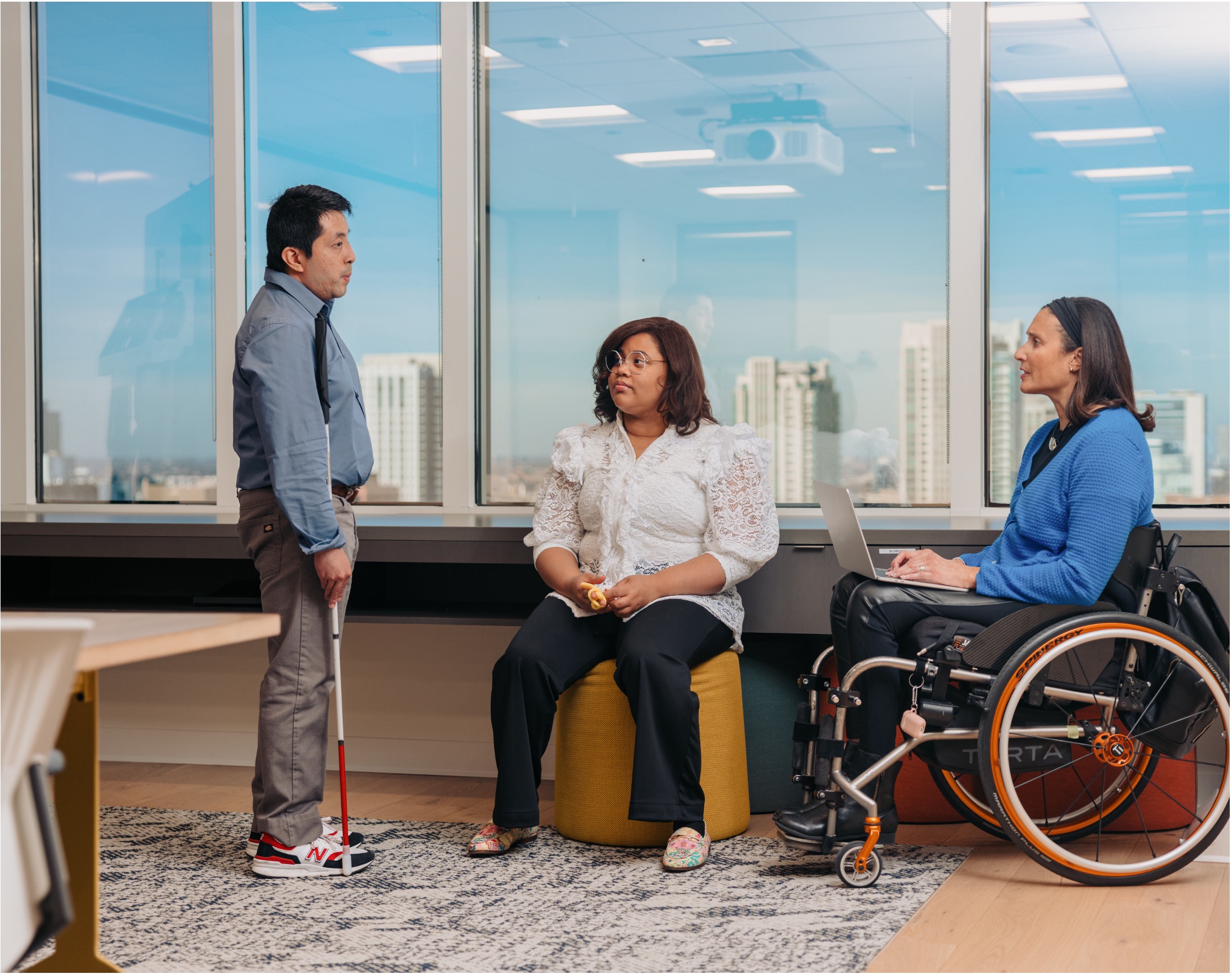 Three people talking in an office. One person is holding a red and white cane, and another is using a wheelchair.